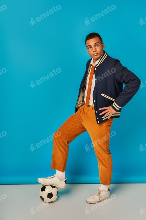Preview: trendy african american student in jacket, orange pants stepping on soccer ball on blue, hand on hip