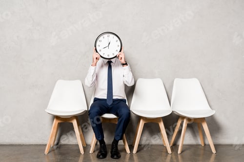Preview: man in formal wear covering face with clock while sitting on chair
