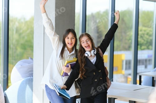 Preview: Two young women with book chatting while standing in college corridor.