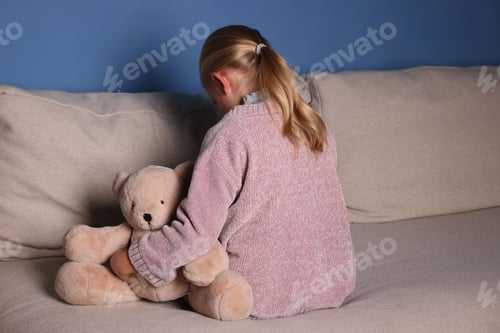 Preview: Sad little girl with toy bear on sofa indoors, back view