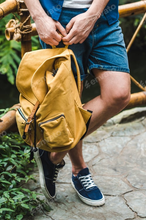 Preview: cropped shot of stylish man with yellow backpack leaning on fence in park