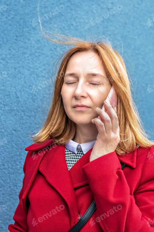 Preview: A pretty young woman is talking on a mobile phone. A serious girl in a coat on a blue background