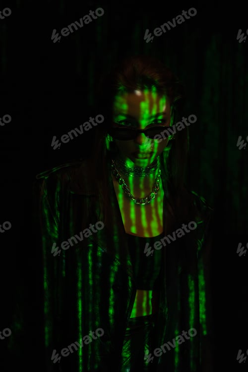 Preview: young woman in trendy sunglasses looking at camera in green neon light isolated on black