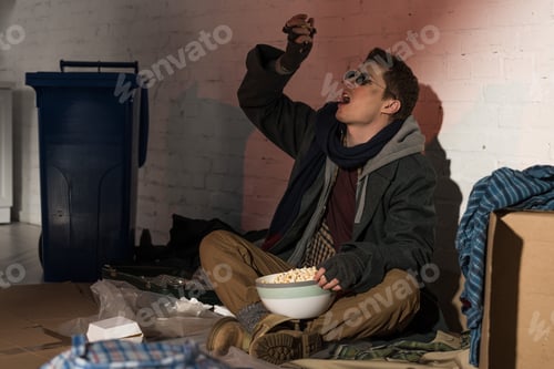 Preview: homeless man eating popcorn while sitting on garbage dump