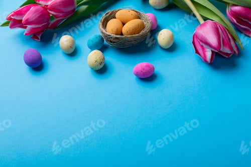 Preview: High angle view of easter candies in small nest with pink tulips on blue background with copy space