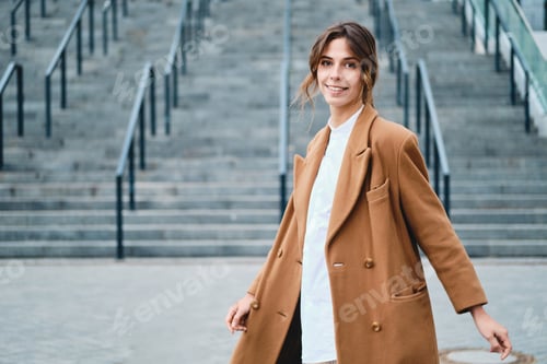 Preview: Young attractive stylish woman in coat happily looking in camera outdoor