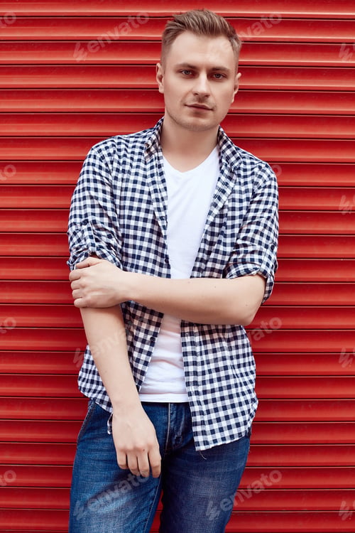 Preview: Portrait Of Young Handsome Man In A Shirt And Jeans Stands Near The Red Metal Gate. Street Style.
