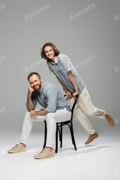 Preview: cheerful teenager leaning on chair next to happy father sitting and smiling on grey