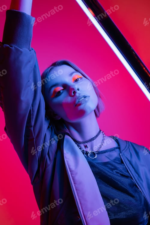 Preview: trendy woman with neon makeup and necklaces holding fluorescent lamp on deep pink and coral
