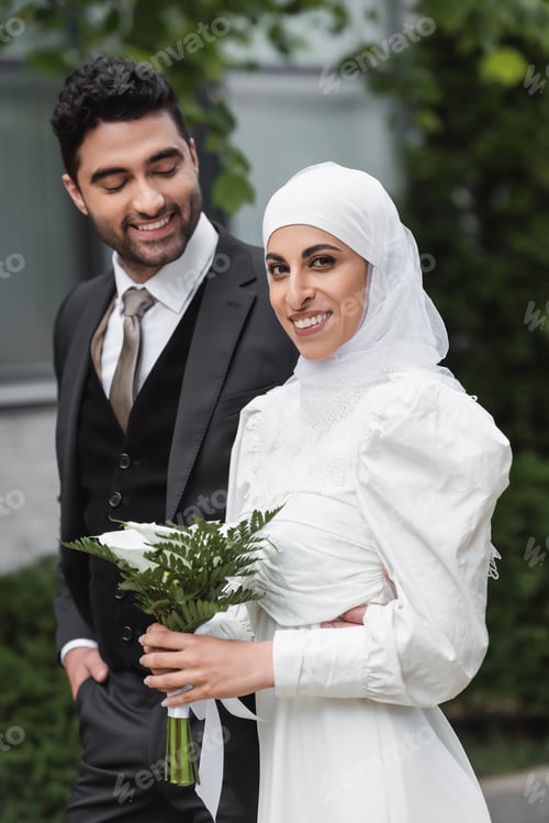 Preview: cheerful groom looking at muslim bride in wedding hijab and white dress holding bouquet