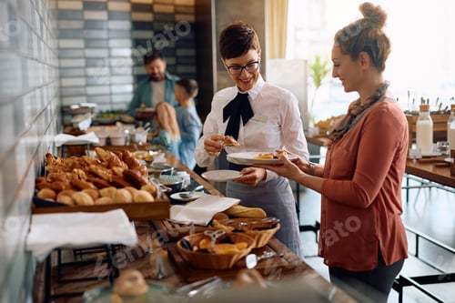 Preview: Happy waitress serving food to hotel guest during buffet breakfast in a restaurant.