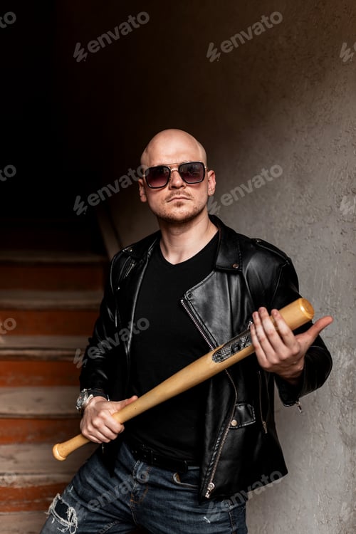 Preview: Brutal bald man in fashion sunglasses in black vintage leather jacket in jeans with baseball