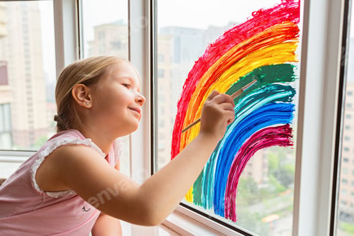 Preview: Happy Caucasian little girl drawing bright and colorful rainbow on window at home