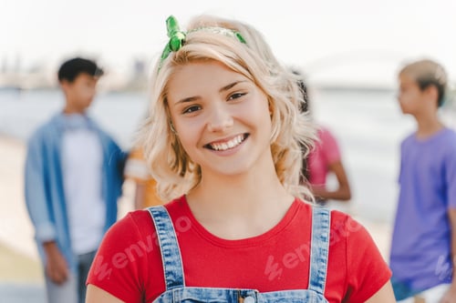 Preview: Smiling teenage girl wearing red t shirt looking at camera on the street with friends on background