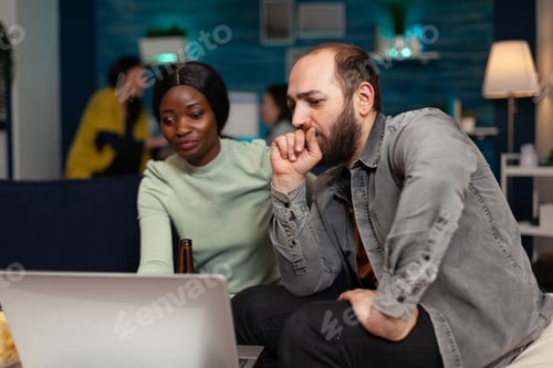 Preview: Multi-ethnic friends sitting on couch watching online video on laptop enjoying spending time
