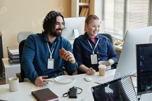 Preview: Two happy young office managers having snacks in front of computer screen