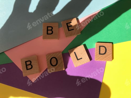Preview: Be Bold in wooden alphabet letters on colourful multicoloured background with shadow of human hand