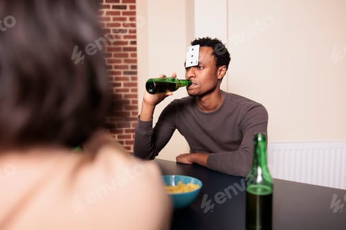 Preview: Confident young adult man with plastic card on forehead drinking beer while playing charades with