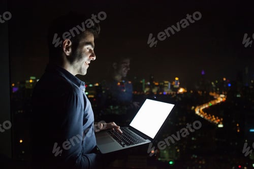 Preview: A young man working on his laptop against the backdrop of a vibrant city night skyline.
