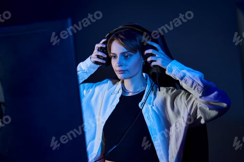 Preview: focused woman wearing headphones and looking at computer in a blue-lit room, cybersport game concept