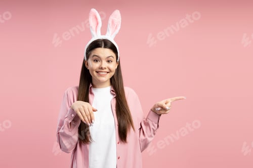 Preview: Young woman wearing bunny ears pointing with finger on pink background