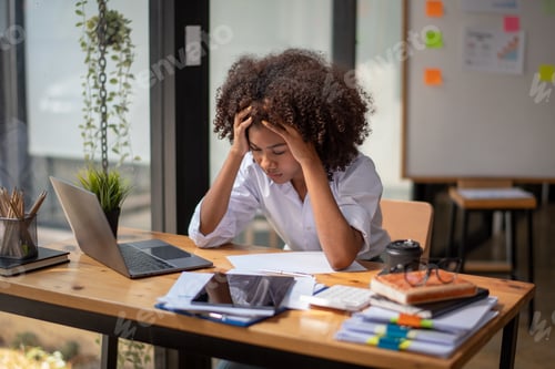 Preview: A woman is sitting at a desk with a laptop and a stack of papers