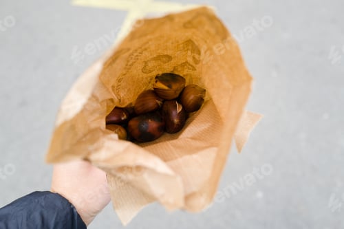 Preview: Hand holding a brown paper bag with warm roasted chestnuts