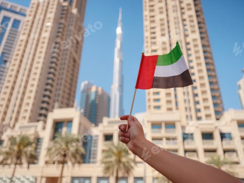 Preview: female hand holding tiny flag of UAE against Dubai skyline.