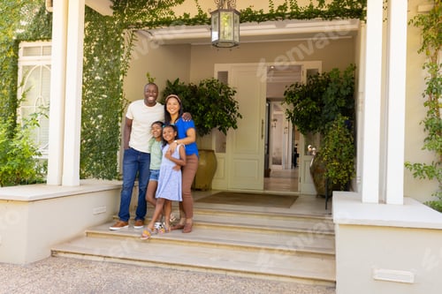 Preview: Smiling family of four with daughters standing together on front porch of their home