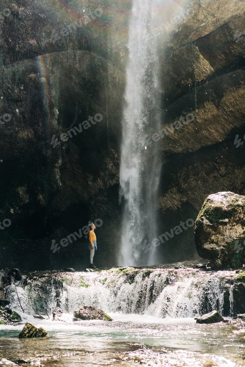 Preview: A tiny figure of a young active female tourist at a large mountain waterfall.