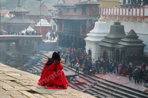 Preview: Back view of woman in red sari watching mournful cremation ceremony at Pashupatinath Temple complex