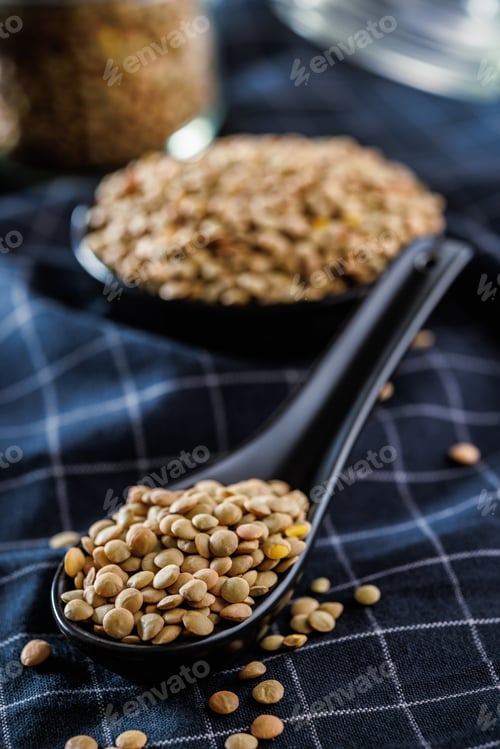 Preview: Uncooked lentil legumes in ceramic spoon on checkered napkin.