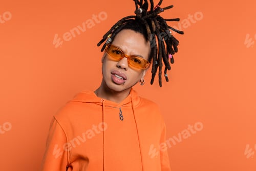 Preview: doubtful multiracial man with dreadlocks and stylish sunglasses looking at camera isolated on coral