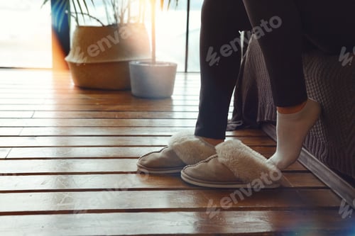 Preview: Woman putting on soft plush warm slippers while sitting on sofa at cozy home in cold season