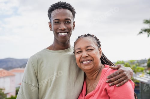 Preview: Happy african mother and adult son hugging each other while smiling on camera at home terrace