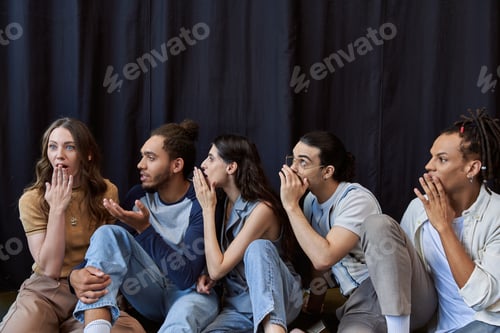 Preview: multiethnic colleagues sitting and telling gossip to shocked woman near black drape in office