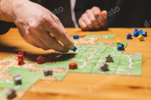 Preview: men friends have fun playing the carcassonne board game at night. male hands and game cards and chip