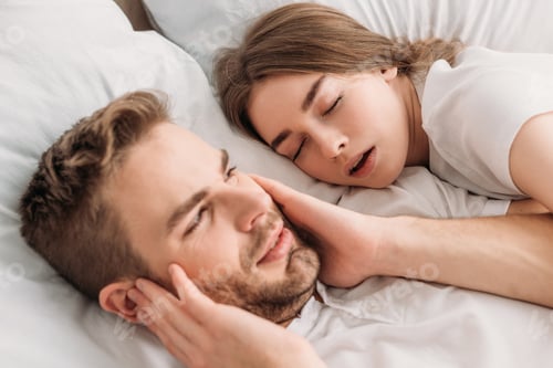 Preview: exhausted man plugging ears with hands while lying in bed near snoring wife