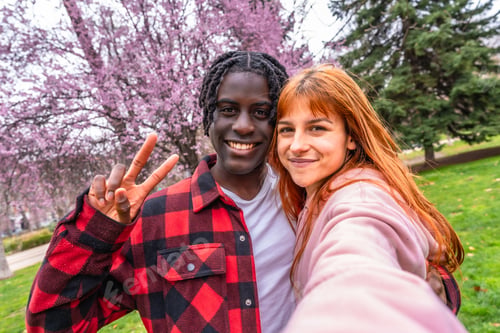 Preview: Happy multiracial couple taking a selfie in a blossoming park making victory sign
