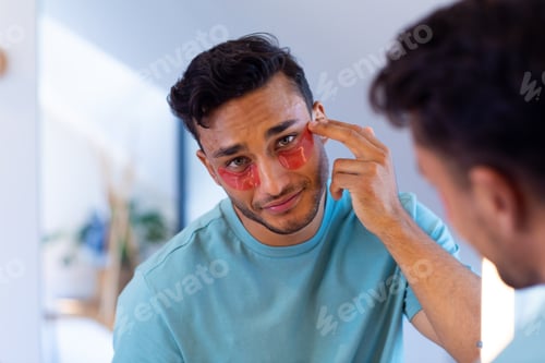 Preview: Biracial man looking in mirror and applying under eye masks in bathroom