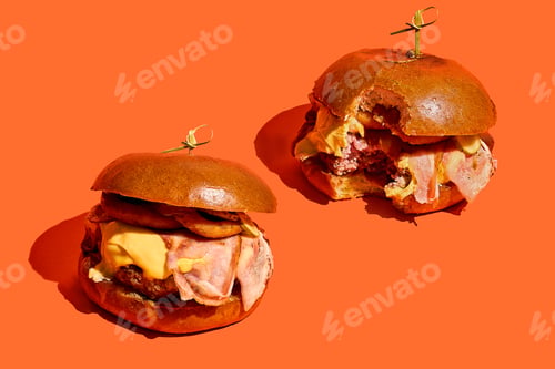 Preview: Two burgers with bacon and onions, melted cheese. One bitten burger on orange background