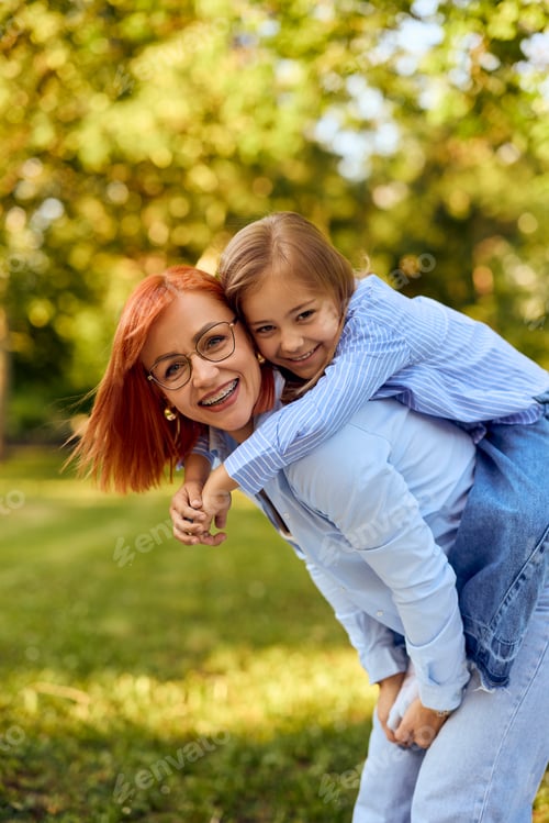 Preview: Portrait of a happy mother and daughter, holding her on the back, posing for the camera in the park.