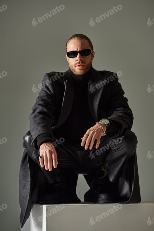 Preview: fashionable male model in trendy sunglasses and black attire sitting on top of on white cube on grey