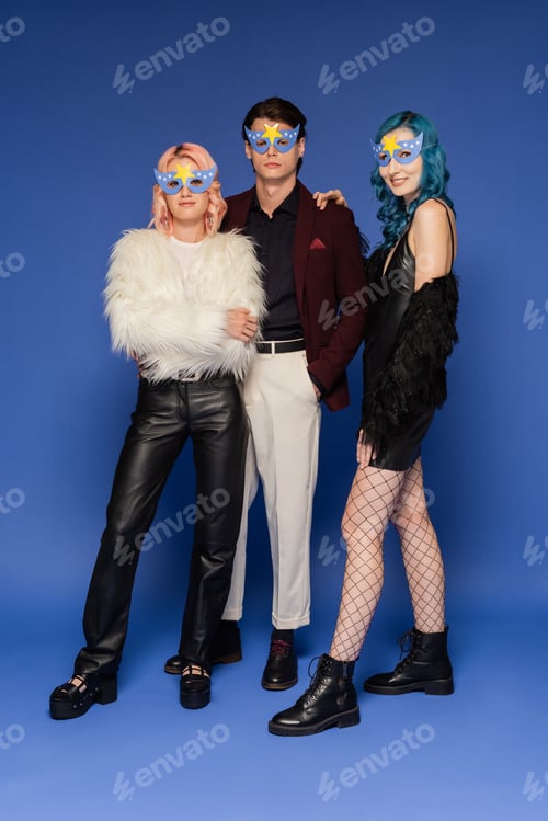 Preview: full length of nonbinary people in fashionable clothes and party masks looking at camera on blue