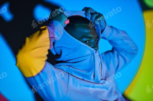Preview: good looking young man posing while putting on hood in digital projector lights, fashion concept