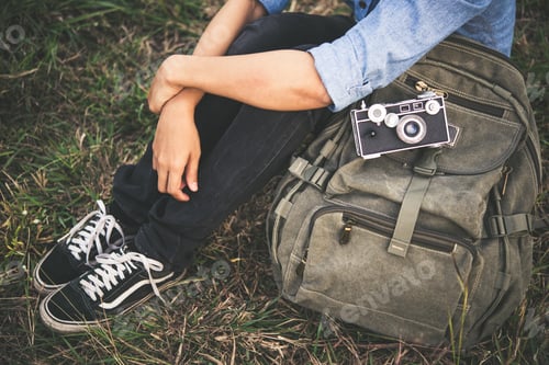 Preview: Young hipster traveller woman with backpack sitting in the grass during sunset.