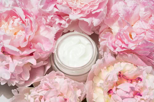 Preview: Open cream jar with pink peonies on soft backdrop. Fresh, floral beauty flat lay for skincare