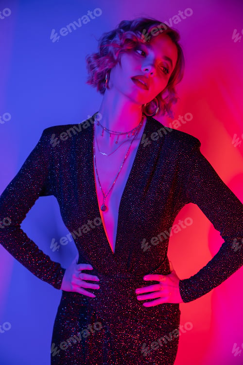 Preview: young woman with blonde hair posing with hands on hips in black tight dress on purple and pink
