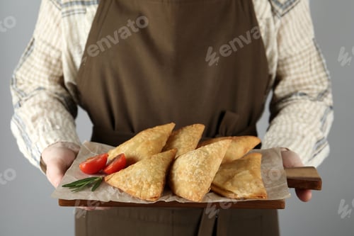 Preview: Woman holding wooden board with delicious samosas on grey background, closeup