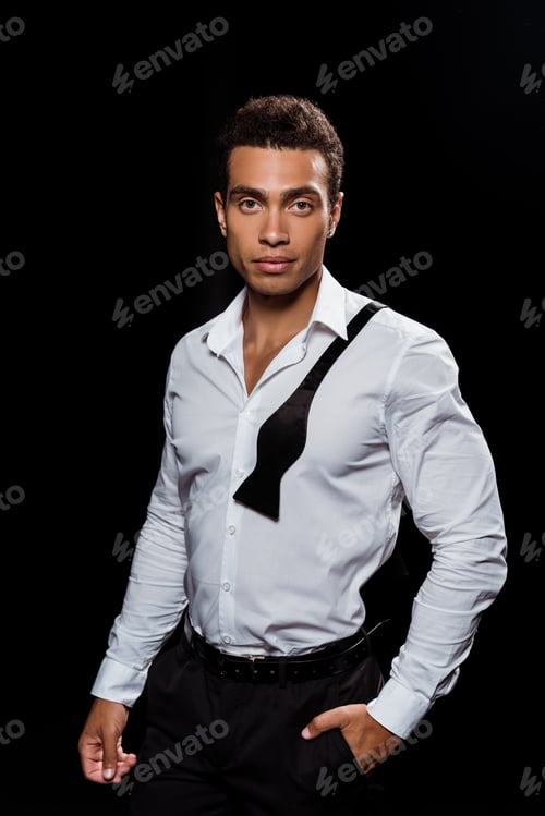 Preview: handsome mixed race man looking at camera while standing with hand in pocket isolated on black